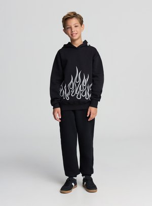 Felpa nera con cappuccio e motivo a fiamma bianca sull'orlo, abbinata a pantaloni della tuta neri e scarpe nere. Il tessuto sembra morbido e casual.