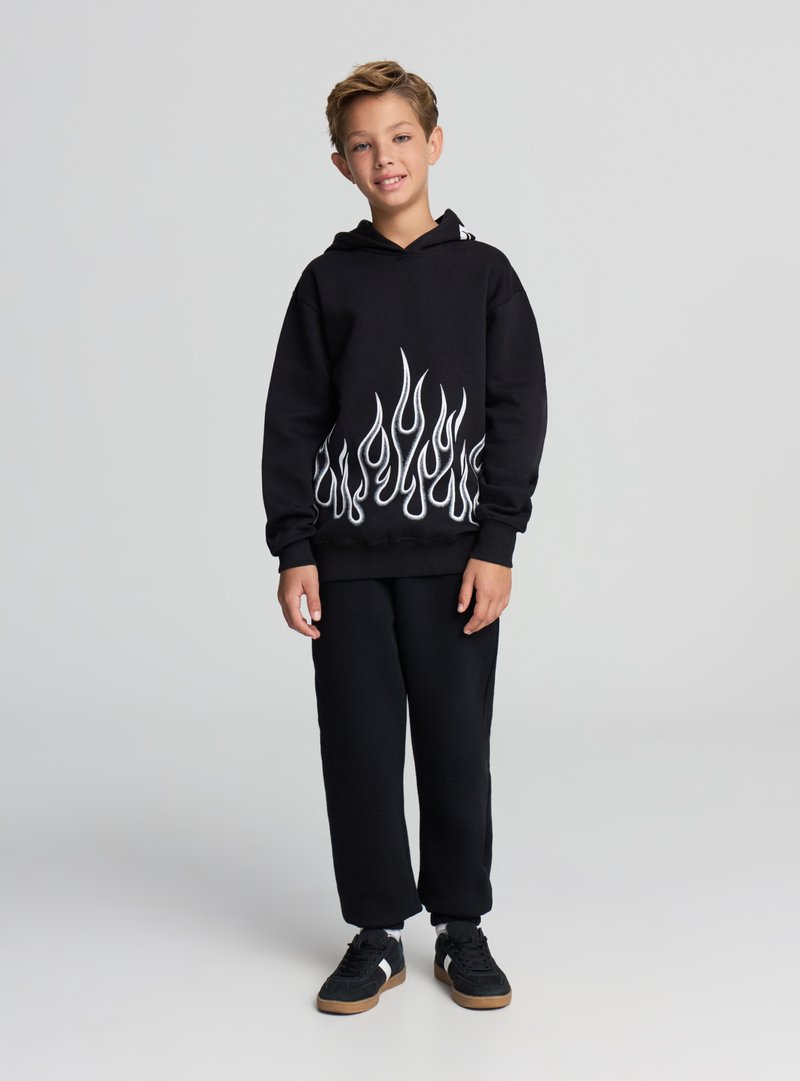 Felpa nera con cappuccio e motivo a fiamma bianca sull'orlo, abbinata a pantaloni della tuta neri e scarpe nere. Il tessuto sembra morbido e casual.