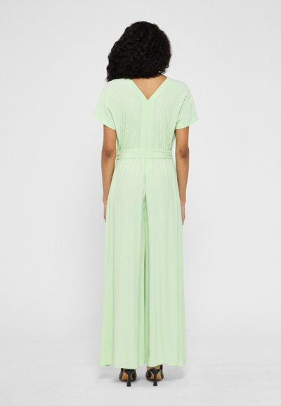 Γυναίκα με σγουρά μαλλιά, φορώντας ένα ανοιχτό πράσινο φαρδύ σορτς με κοντά μανίκια και μαύρες ψηλοτάκουνες σανδάλια, γυρισμένη πλάτη.