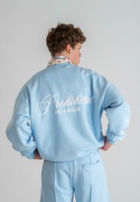 Helles blaues Sweatshirt mit weißem geschwungenem Text "Prohibited 10119 BERLIN" auf dem Rücken, kombiniert mit passenden hellblauen Hosen. Glatte Textur.