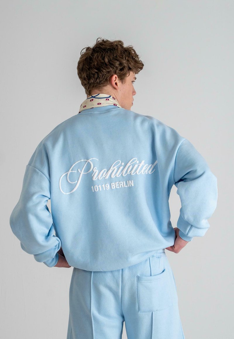 Helles blaues Sweatshirt mit weißem geschwungenem Text "Prohibited 10119 BERLIN" auf dem Rücken, kombiniert mit passenden hellblauen Hosen. Glatte Textur.