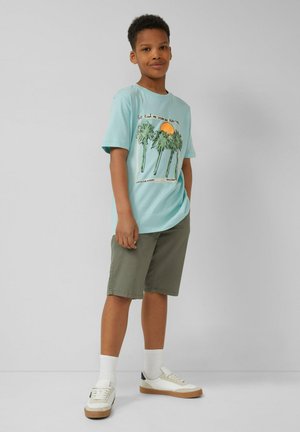 Jongen staat, draagt een lichtblauw T-shirt met palmboom en zonsondergang grafiek, olijfgroene shorts, witte sokken en witte sneakers.