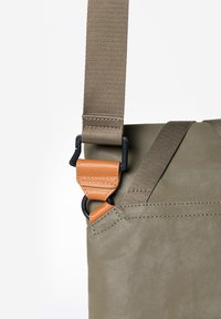 Olijfgroene tas met een gestructureerde afwerking, verstelbare stoffen riem en oranje lederen accenten bij het beslag voor extra duurzaamheid.