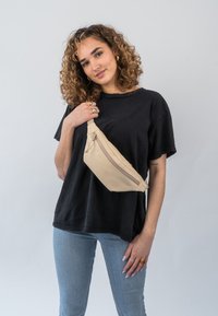 Barna övtáska puha anyagból, cipzárral záródik. Slejkes és áramvonalas formája van, és egy fekete póló fölött a mellkasra van felakasztva.