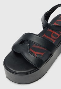 Svarte platåsandaler med tre brede stropper og en definert bølgekutt. Har en teksturert svart foten med rødt logo-print.