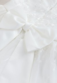 Grand nœud en satin blanc noué sur un tissu en dentelle blanche avec broderie florale, partie d'un vêtement ou accessoire délicat.