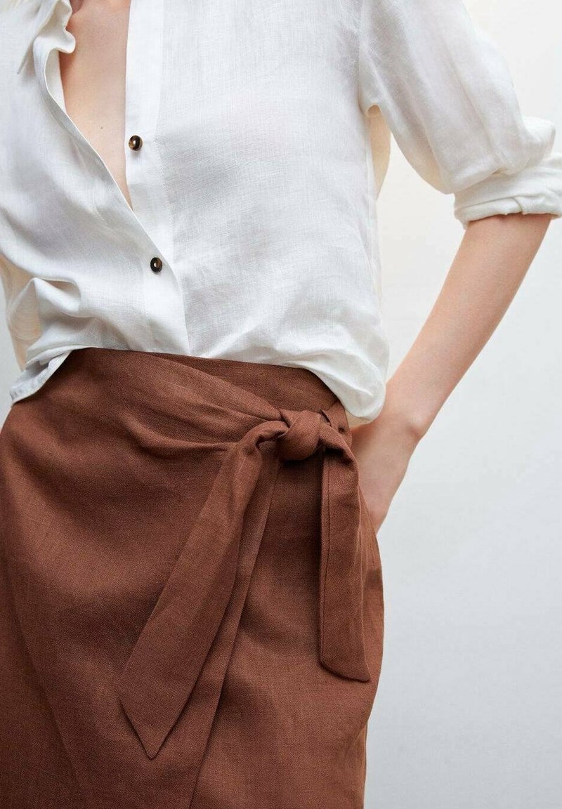 Chemise blanche boutonnée avec manches retroussées associée à une jupe portefeuille marron taille haute nouée en un nœud sur le côté.