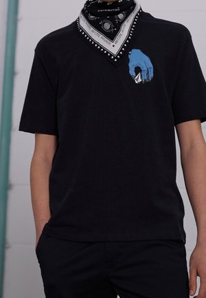 Osoba w czarnej koszulce z niebieskim nadrukiem dłoni trzyma kopertę na klatce piersiowej oraz czarną bandanę z białymi wzorami na szyi.