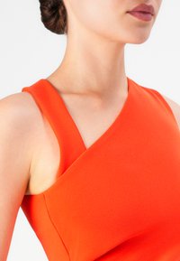 Frau in einem leuchtend orangefarbenen, ärmellosen Kleid mit einem einzigartigen asymmetrischen Ausschnitt und Lippen in einem gedämpften Mauve-Farbton.