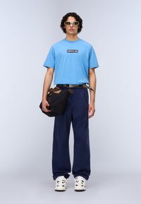 Ανοιχτό μπλε βαμβακερό t-shirt με μαύρο λογότυπο, σκούρο μπλε παντελόνι φαρδιές γραμμές, μαύρη τσάντα ώμου, μεγάλα λευκά αθλητικά παπούτσια και ορθογώνια γυαλιά ηλίου.