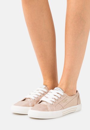 Piernas con zapatillas bajas de color rosa claro con cordones y suelas blancas, de pie sobre una superficie blanca frente a un fondo liso.