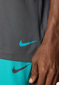 Graues Sportshirt mit türkisfarbenem Nike-Logo. Nahaufnahme des unteren Saums, die eine glatte Textur und kontrastierende Farben zeigt.