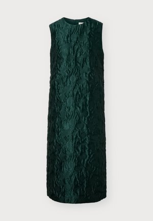 Femme debout portant une robe texturée vert foncé sans manches, longueur genou, et des chaussures plates noires sur un fond clair uni.