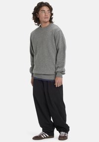 Grauer Strickpullover mit Rundhalsausschnitt, gerippten Bündchen und Saum, getragen über einem blauen Hemd. Weite schwarze Hosen und braune Sneakers runden das Outfit ab.