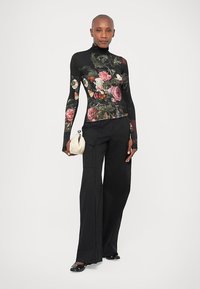 Langermet turtleneck-genser med blomstermønster i sterke farger på svart bakgrunn; kombinert med vide svarte bukser og en kremfarget clutch.