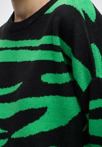 Gros plan d'un pull noir avec un motif rayé abstrait vert porté sur le haut du corps et le cou d'une personne.