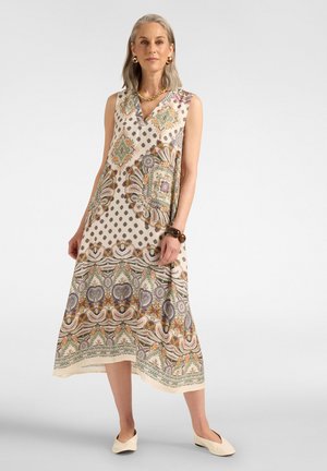 Femme aux cheveux gris portant une robe midi sans manches à motifs géométriques et floraux, des ballerines beige, des boucles d'oreilles en or et un bracelet.