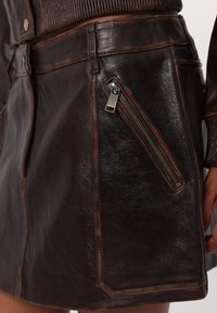 Gros plan sur une jupe en cuir marron foncé avec une poche zippée diagonale et des coutures décoratives le long des coutures.