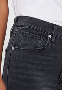 Jeans de mezclilla negra con una cintura de altura media, que presentan un pequeño bolsillo frontal y herrajes en plata. La tela tiene una apariencia suave y texturizada.
