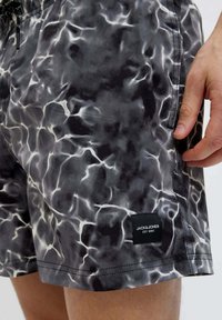 Costumi da bagno neri e grigi con un motivo ondulato, simile all'acqua, vita elastica e un'etichetta di marca sul lato. Tessuto morbido al tatto.