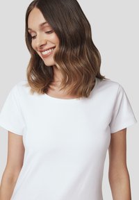Biała koszulka z krótkim rękawem, wykonana z miękkiego materiału, z okrągłym dekoltem i dopasowanym krojem, o gładkiej fakturze i bez widocznych wzorów.
