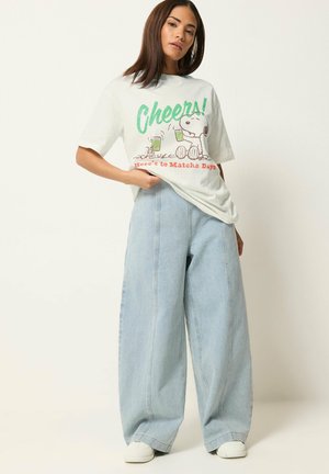 Młoda kobieta ubrana w kremowy T-shirt z grafiką Snoopy i szerokie jasnoniebieskie dżinsy, stojąca na jednolitym tle.