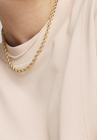 Collana a catena intrecciata in oro stratificata con un delicato filo di perle, appoggiata su uno sfondo di tessuto rosa chiaro con una finitura testurizzata.