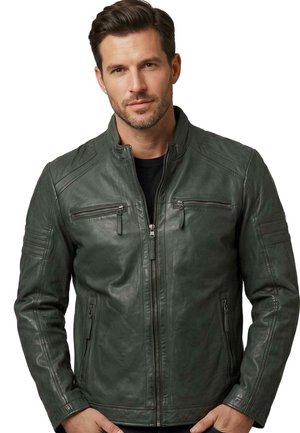 Mann trägt eine dunkelgrüne Lederjacke mit Reißverschlusstaschen auf Brust und Seiten, steht vor weißem Hintergrund.