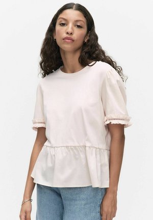 Henkilö, jolla on pitkät kiharat hiukset, pukeutuneena vaaleanpunaiseen röyhelöiseen lyhythihaiseen paitaan ja vaaleansinisiin farkkuihin, seisoo yksiväristä valkoista taustaa vasten.