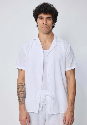 Homme aux cheveux bouclés portant une chemise blanche ouverte à manches courtes sur un débardeur blanc et un pantalon à cordon, avec un tatouage tribal sur l'avant-bras gauche.