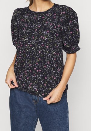 Vrouw draagt een zwarte korte mouwen blouse met kleine roze, paarse en groene bloemmotieven, gecombineerd met blauwe spijkerbroek.