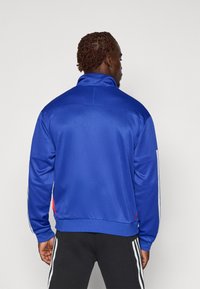 Blå idrettsjakke med høy krage, glatt tekstur og hvite stripemerker på ermene. Rødt logodetalj ved kanten.