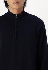 Maglione con mezza zip di colore blu navy realizzato in un materiale lavorato a maglia. Presenta un colletto a costine e un design con mezza zip con dettagli di branding minimi.