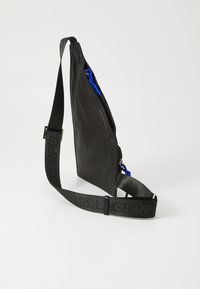 Svart läder crossbody-väska med en slank design. Har en blå dragkedja och ett texturerat svart band med varumärkesnamnet präglat.
