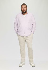 Lichtpaarse lange mouwen shirt, beige broek en witte sneakers. Het shirt heeft een voorzak. Het model staat met de handen in de zakken.