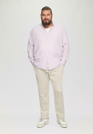 Lichtpaarse lange mouwen shirt, beige broek en witte sneakers. Het shirt heeft een voorzak. Het model staat met de handen in de zakken.