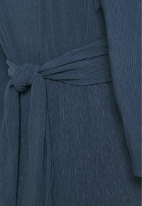 Ciemnoniebieska sukienka o teksturowanej powierzchni z węzłowym paskiem materiałowym. Materiał posiada pionowe fale, podkreślające płynny krój i miękką fakturę.