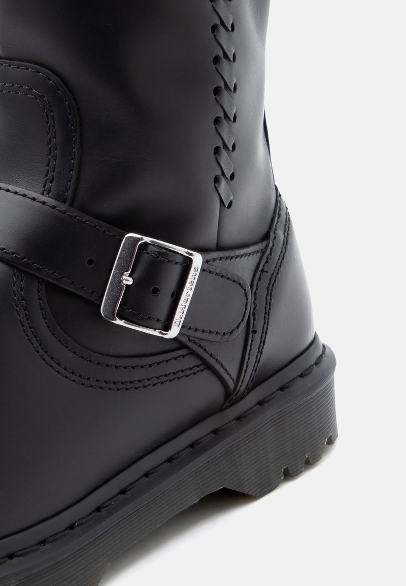 Schwarzer Lederstiefel mit seitlichem Schnallenriemen, genähten Details und gerippter Gummisohle, vor einem weißen Hintergrund dargestellt.