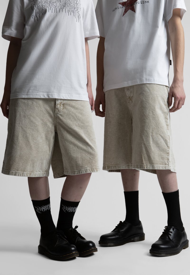 Δύο άτομα φοράνε λευκά T-shirts με σχέδια, μπεζ βερμούδες από κορδόνι μέχρι το γόνατο, μαύρες κάλτσες μέσης και μαύρα δερμάτινα παπούτσια, στέκονται δίπλα-δίπλα.