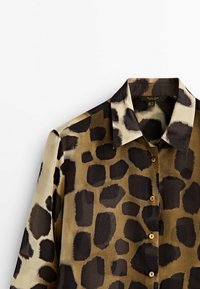 Chemise à manches longues avec boutons, ornée de grandes taches irrégulières noires et brun foncé sur un fond tan, dotée d'un col pointu.