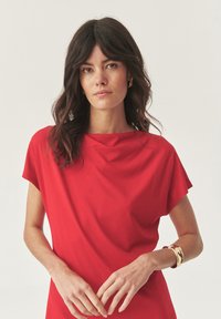 Robe rouge à manches courtes avec un décolleté drapé, en tissu lisse. Présente une coupe plate et décontractée ainsi qu'un bracelet métallique doré au poignet.
