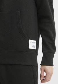 Sweatshirt noir avec un ourlet et des poignets côtelés, présentant une poche avant et une étiquette tissée sur le côté avec des détails textuels. Texture douce.