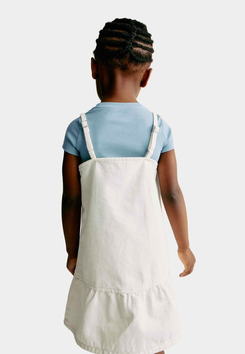 Hvid bomuldsdungaree-kjole med en flæsekant, båret over en lysblå ribbet kortærmet top. Bagsiden viser justerbare stropper.