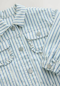Veste en denim rayée bleu clair et blanche avec un col, deux poches poitrine, des boutons pression en argent et des bords de poches volantés.