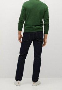 Homme portant un pull vert à manches longues, un jean bleu foncé et des baskets blanches, debout de dos devant un fond uni.