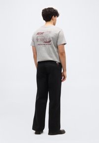 Άνδρας που φοράει γκρι μπλούζα με κόκκινη εκτύπωση με χάμπουργκερ, πατάτες και ποτό και μαύρο παντελόνι, στέκεται γυρισμένος με την πλάτη σε μονόχρωμο φόντο.