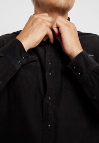 Camisa de pana negra con botones de presión, bolsillos en el pecho, textura acanalada y una pequeña etiqueta de logo en el cuello. Se ven manos ajustando el cuello.