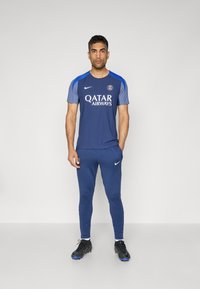 Marine atletisk sæt fra Nike, der har en kortærmet top med blå detaljer, logo og Qatar Airways tekst, parret med matchende tætsiddende bukser.