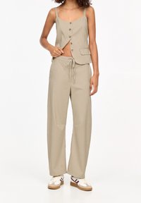 Femme portant un gilet sans manches beige boutonné et un pantalon large assorti avec des baskets blanches, posant avec la main ajustant le gilet.