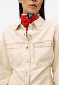 Frau trägt eine beigen Jeansjacke mit Fronttaschen und einem rot gemusterten Halstuch, das zu einem Knoten gebunden ist.